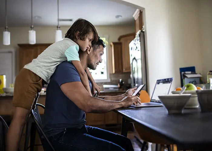 Dad at table working on a laptop boy on his back