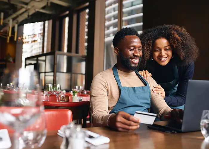 Business owner holding credit card and using laptop while seated with woman leaning over and smiling next to him