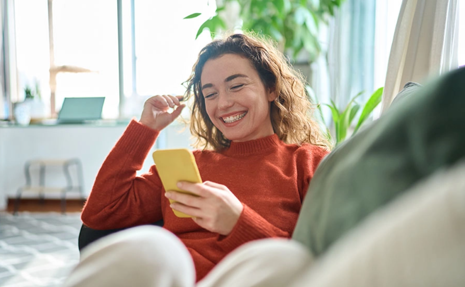 Young woman sitting on the couch and smiling while using her smartphone